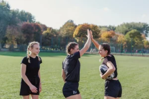 Tres jóvenes jugadoras de rugby celebran en un campo verde, una de ellas sostiene un balón de rugby mientras dos se dan un choque de manos, reflejando camaradería y entusiasmo por el deporte.