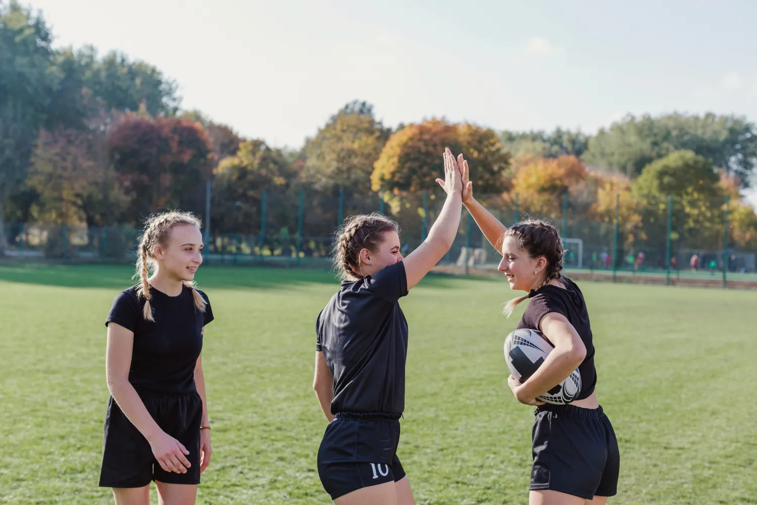 Tres jóvenes jugadoras de rugby celebran en un campo verde, una de ellas sostiene un balón de rugby mientras dos se dan un choque de manos, reflejando camaradería y entusiasmo por el deporte.