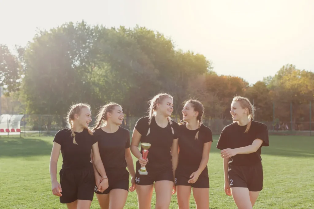 Grupo de cinco chicas sonrientes en un campo de fútbol, vestidas con camisetas y pantalones cortos negros, celebrando con un trofeo en manos. La escena muestra un ambiente alegre y amistoso, con árboles y luz natural de fondo.