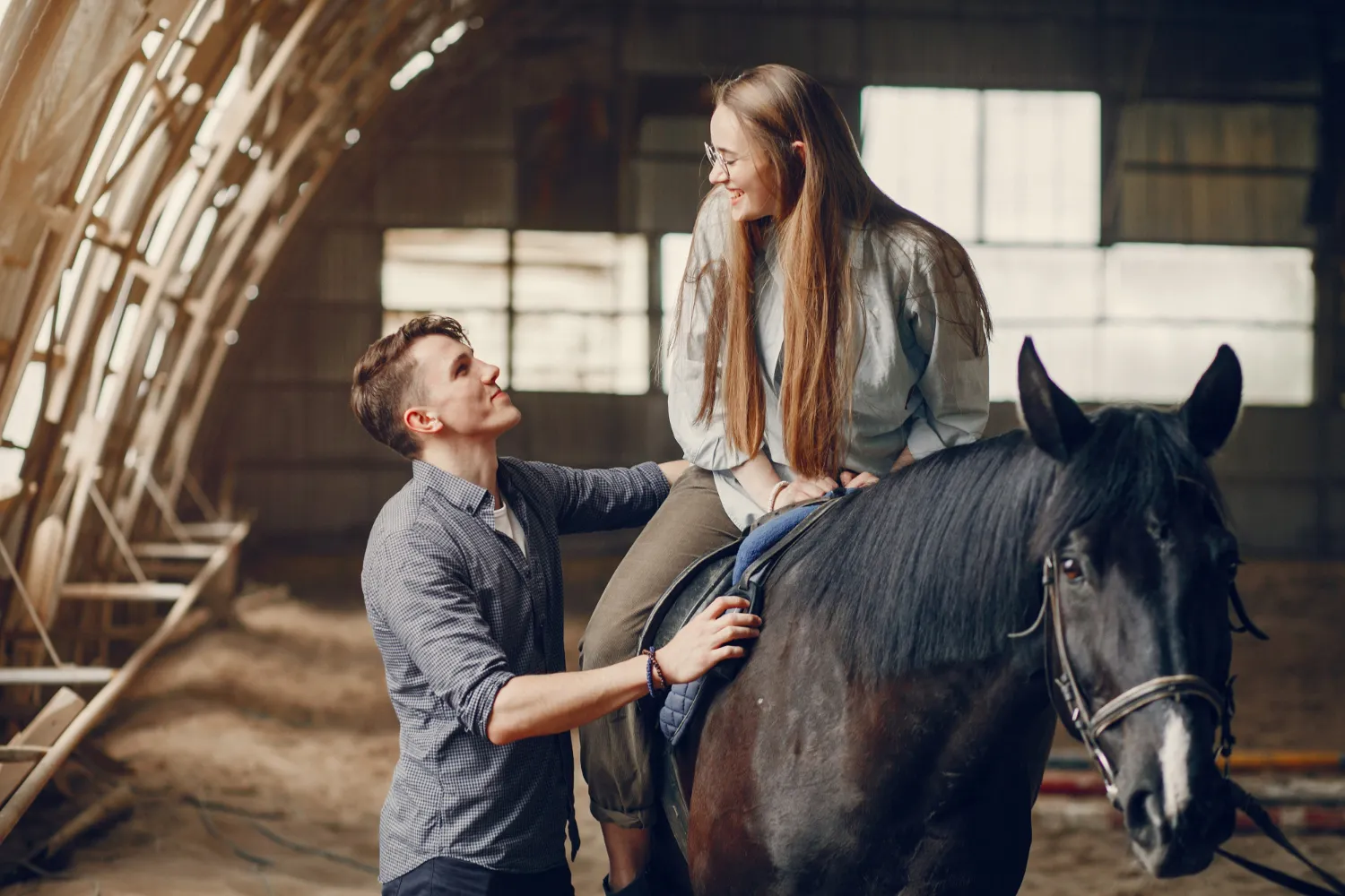Pareja sonriente en un establo, el hombre ayuda a la mujer a montar un caballo negro, creando un ambiente de conexión y amor por los animales.