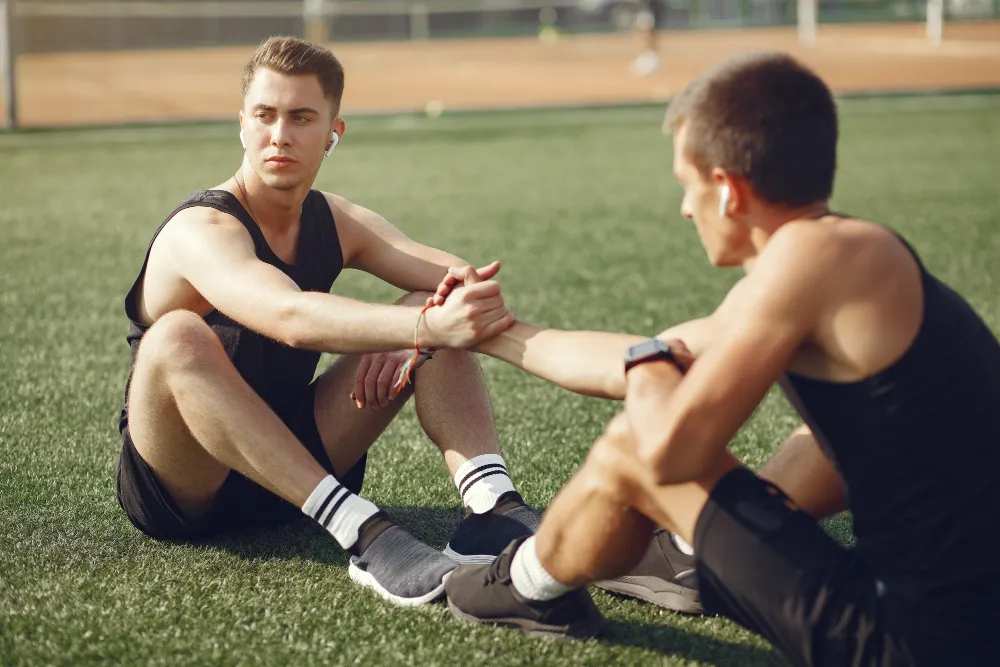 Dos jóvenes haciendo ejercicio en un campo de hierba, sentados en el suelo mientras compiten en un juego de fuerza con las manos. Ambos llevan ropa deportiva y auriculares, mostrando un ambiente de entrenamiento y camaradería.