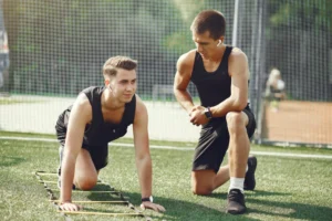 Dos jóvenes entrenando en un campo de césped artificial, uno en posición de ejercicio sobre una escalera de agilidad y el otro observando y dando instrucciones. Ambos visten ropa deportiva y se enfocan en mejorar su rendimiento físico.