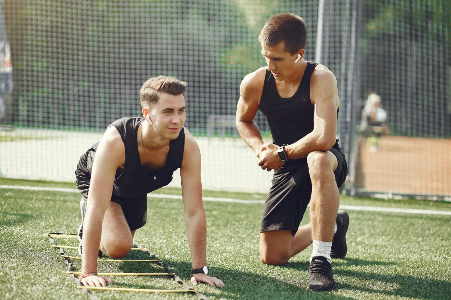 Dos jóvenes entrenando en un campo de césped artificial, uno en posición de ejercicio sobre una escalera de agilidad y el otro observando y dando instrucciones. Ambos visten ropa deportiva y se enfocan en mejorar su rendimiento físico.